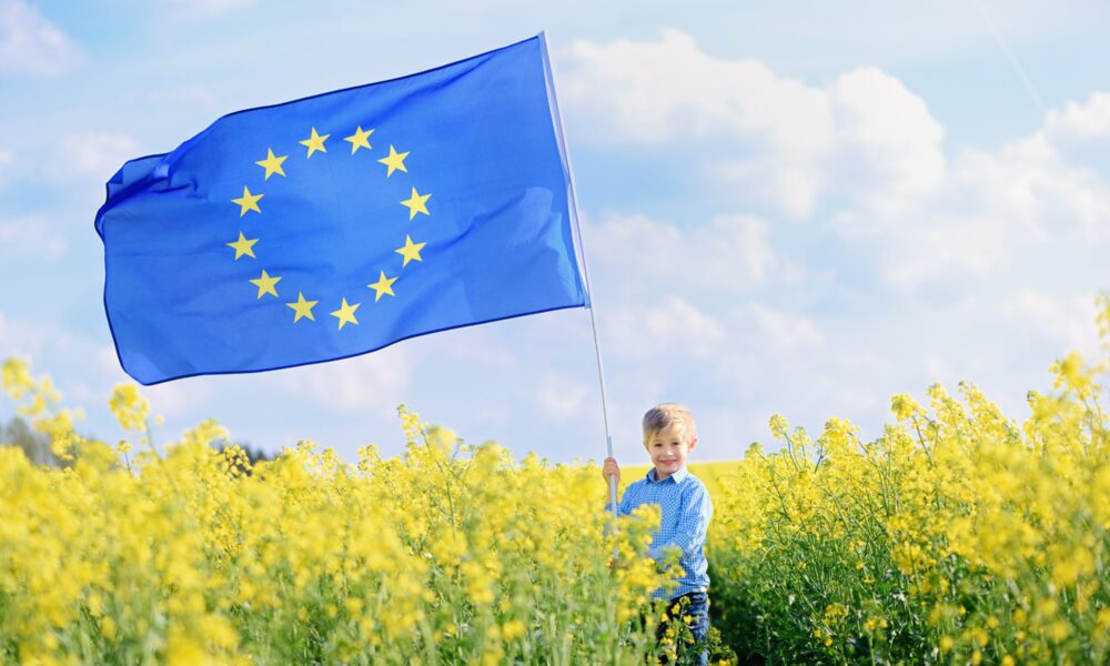 Ein Junge steht in einem gelben Rapsfeld. Er hält eine große Europa-Flagge. Diese weht im Wind