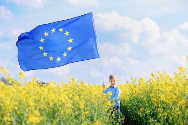 Ein Junge steht in einem gelben Rapsfeld. Er hält eine große Europa-Flagge. Diese weht im Wind