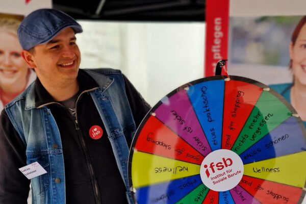 Ein junger Mann steht hinter einem bunten Glücksrad. In der Mitte das Logo IfSB