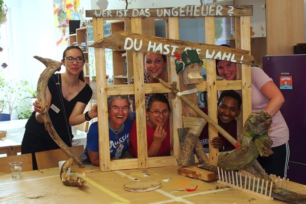 Fünf junge Menschen schauen durch die Zwischenräume einer Holzpalette. Eine junge Frau steht daneben. Auf der Palette steht: Wer ist hier das Ungeheuer. Du hast die Wahl.