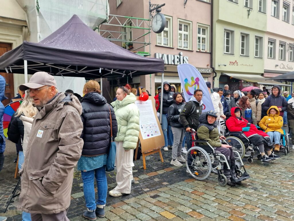 Das Bild zeigt viele Menschen. Manche Menschen haben eine Behinderung und fahren mit dem Rollstuhl. Sie unterhalten sich.
