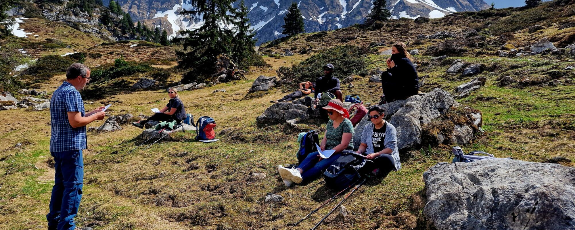 Eine Gruppe junger Menschen sitzt auf einer Wiese vor einer Bergkulisse. Ein Mann steht vor ihnen und liest aus einem Text vor.