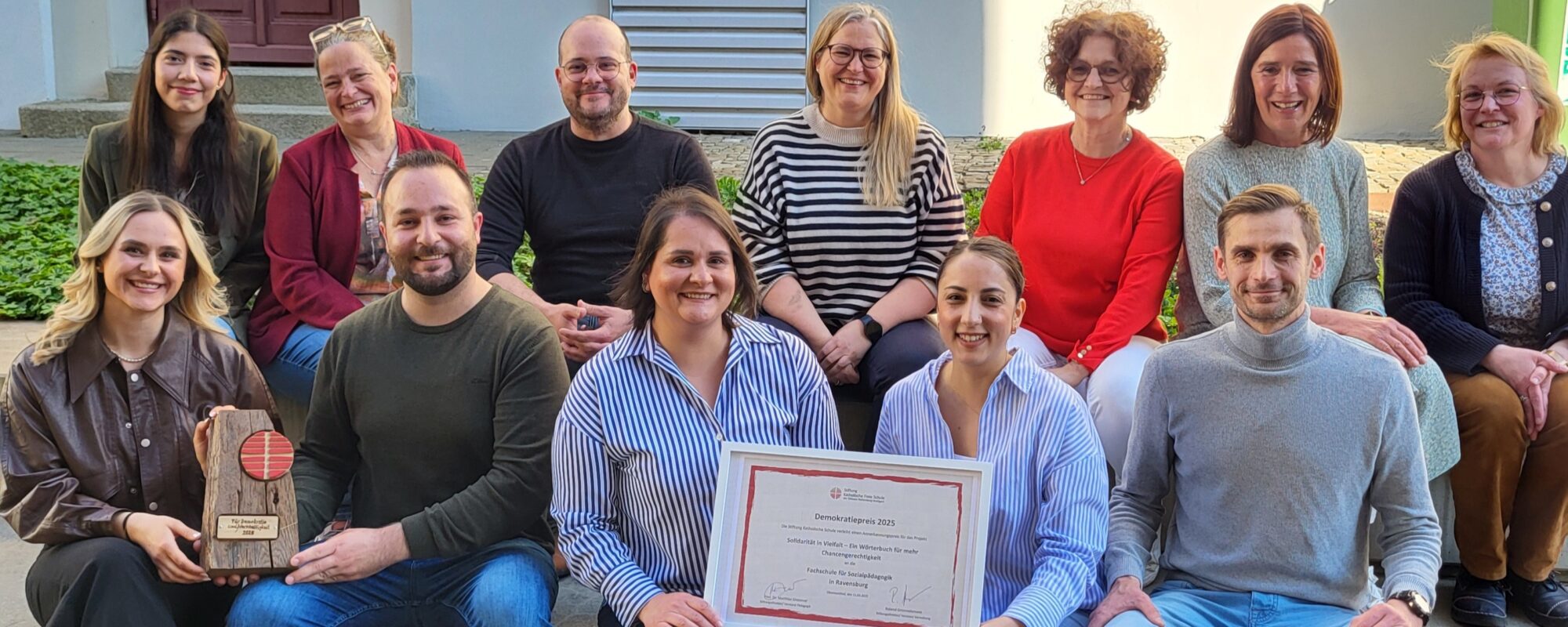Das Bild zeigt eine Gruppe von Personen. Lehrende und Auszubildende der Fachschule Sozialpädagogik. Zwei Personen halten eine Urkunde mit der Aufschrift Demokratiepreis 2025. Demokratie in Vielfalt ein Wörterbuch für mehr Chancengerechtigkeit