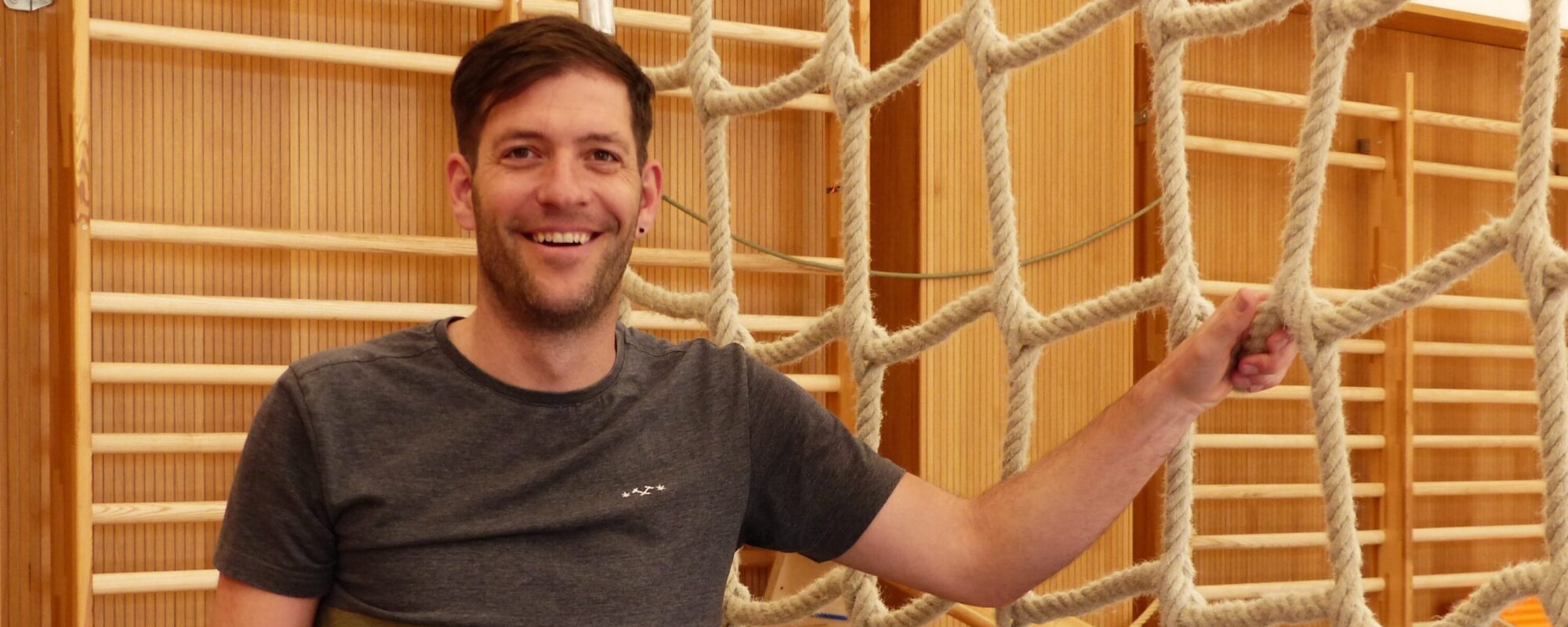 Das Bild zeigt Raphael Frirdich, einen Lehrer für Psychomotorik am IfSB. Er steht im Sportraum vor einem Kletternetz