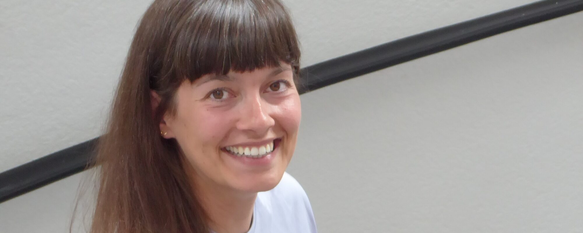 Sabine Schorpp, eine Frau Mitte dreißig, lange braune Haate und weißes Shirt. Sie hält sich mit der Hand am Handlauf einer Treffe und lacht in die Kamera