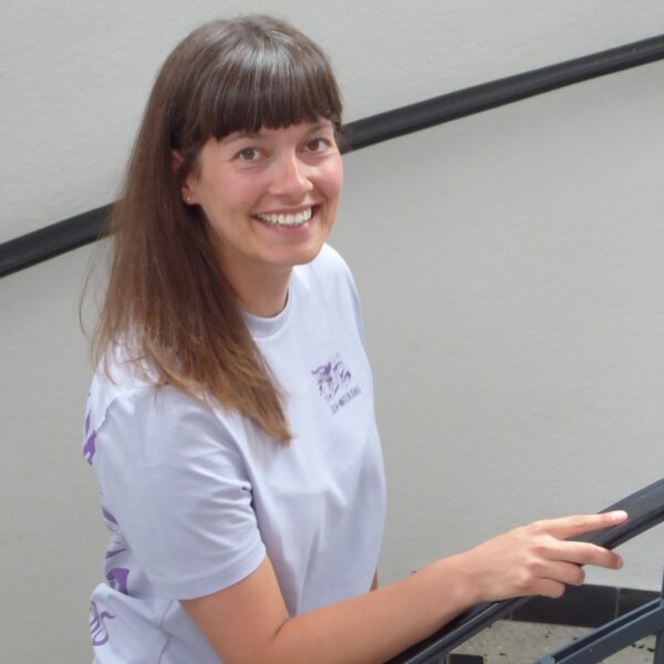 Sabine Schorpp, eine Frau Mitte dreißig, lange braune Haate und weißes Shirt. Sie hält sich mit der Hand am Handlauf einer Treffe und lacht in die Kamera