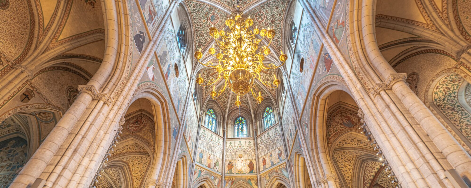 Das Foto zeigt die Sankt Patrick Kathedrale in New York von innen. Der Blick wird durch die hohen Säulen nach oben gezogen. Unter der Decke hängt ein prächtiger goldener Leuchter