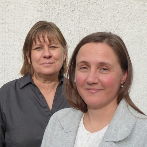 Das Foto zeigt die beiden Religionspädagoginnen Heli Pfizenmaier und Daniela Alker. Frau Pfizenmaier hat halblanges Haar und trägt eine dunkelgraue Bluse. Frau Alker hat langes Haar, trägt einen Mittelscheitel und ist mit einem grauen Blazer und weißen Shirt bekleidet. Beide sind Damen mittleren Alters.