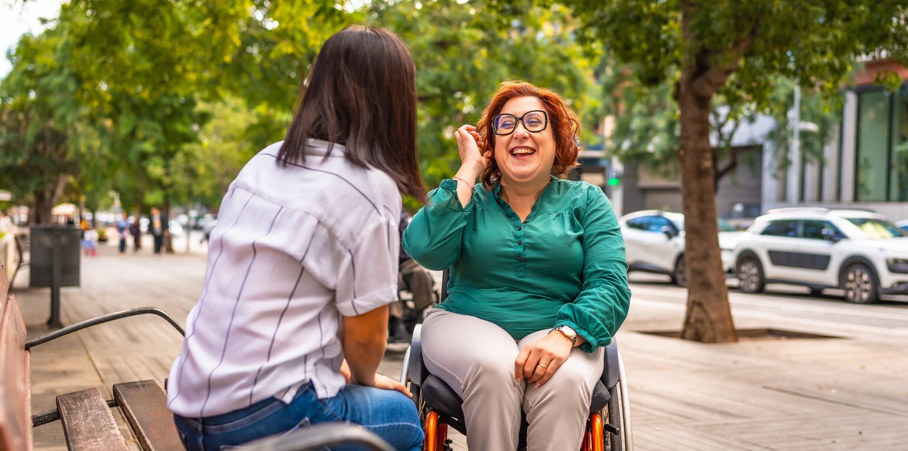 Eine Frau im Rollstuhl mit grünem Oberteil spricht mit einer sitzenden Heilerziehungsassistentin mit weißem Hemd auf einer Parkbank in einer Baumallee.