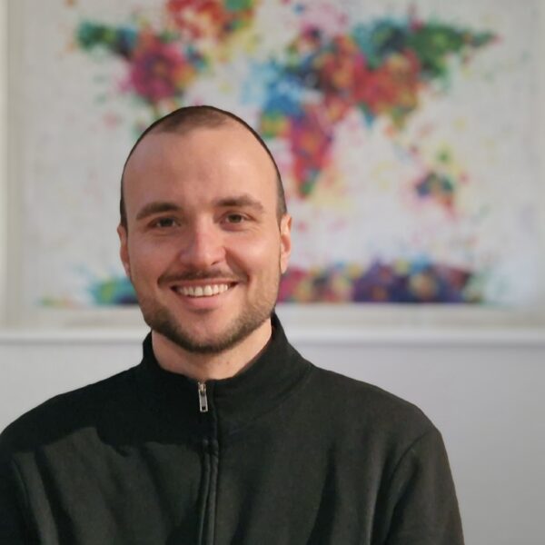 Auf dem Foto ist ein Portraitbild von Jonas Ruf abgebildet. Der junge Mann ist schwarz gekleidet und lächelt in die Kamera. Im Hintergrund sieht man eine Weltkarte, die in vielen bunten Farben gestaltet ist.