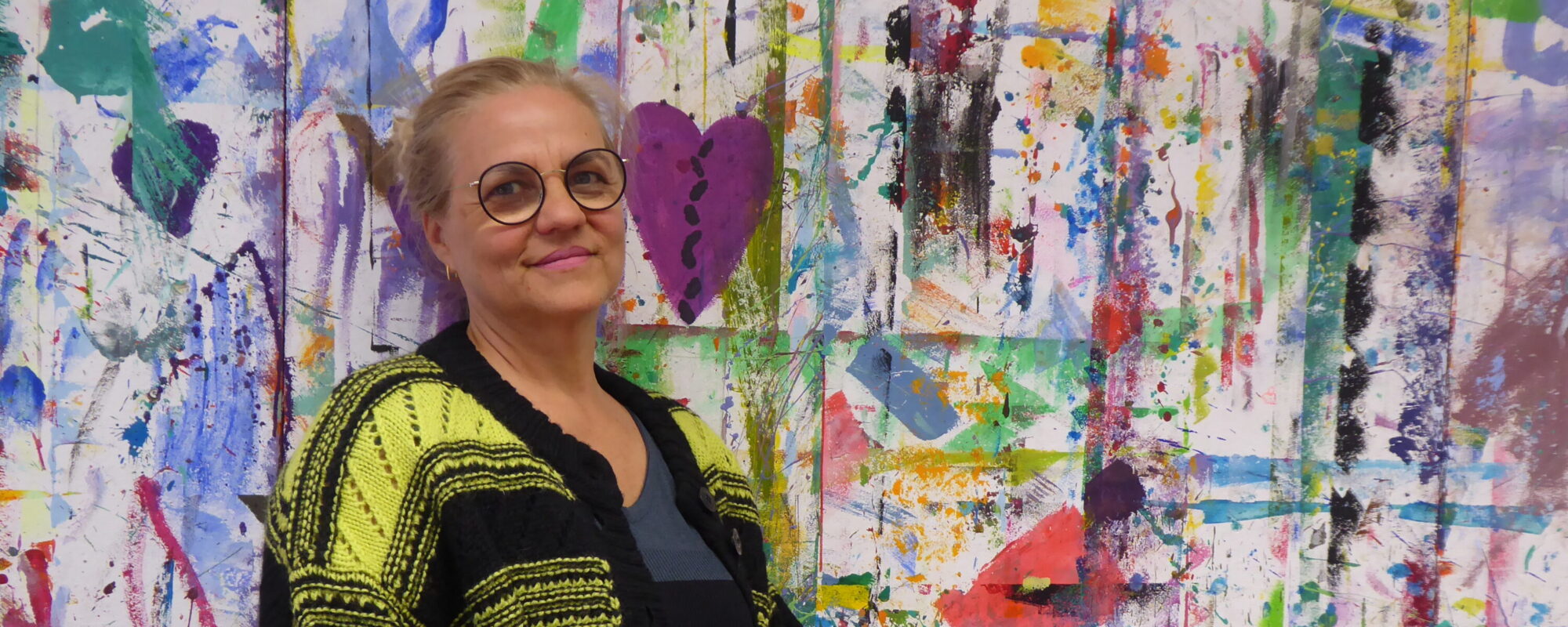 Das Bild zeigt die Kunst-Dozentin Isabella Moser. Sie steht vor einer bunten Malwand, Direkt neben ihr ein lilafarbenes Herz. Isabella Moser ist bekleidet mit einer langen schwarz-gelben Strickjacke. Sie hat ihre Haare streng zurückgekämmt und zusammengebunden und trägt eine Brille