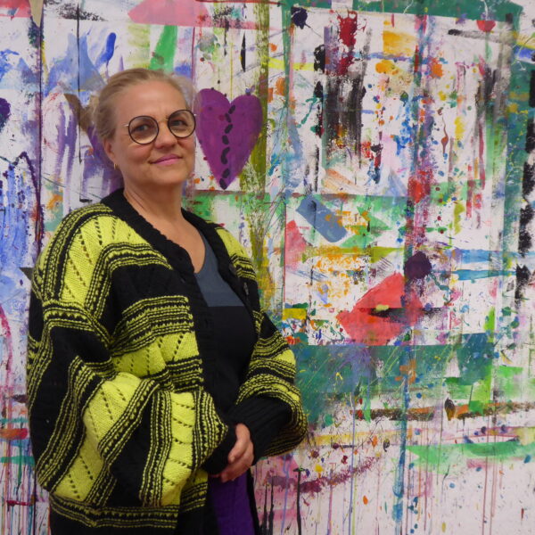 Das Bild zeigt die Kunst-Dozentin Isabella Moser. Sie steht vor einer bunten Malwand, Direkt neben ihr ein lilafarbenes Herz. Isabella Moser ist bekleidet mit einer langen schwarz-gelben Strickjacke. Sie hat ihre Haare streng zurückgekämmt und zusammengebunden und trägt eine Brille